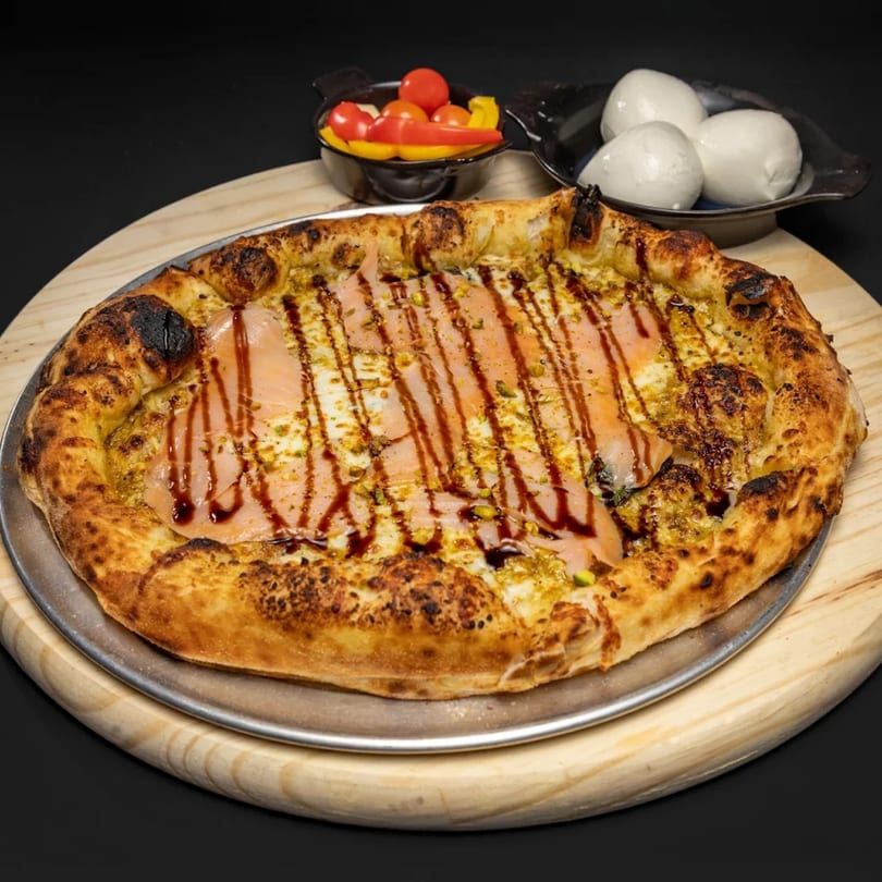 Pizza with pesto, salmon, balsamic glaze, and pistachios, served with cherry tomatoes and mozzarella.