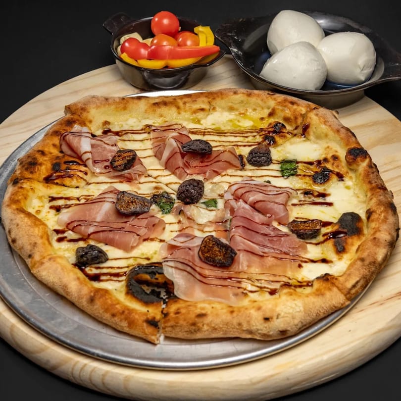 A pizza with prosciutto, figs, and balsamic glaze, alongside bowls of cherry tomatoes and mozzarella.