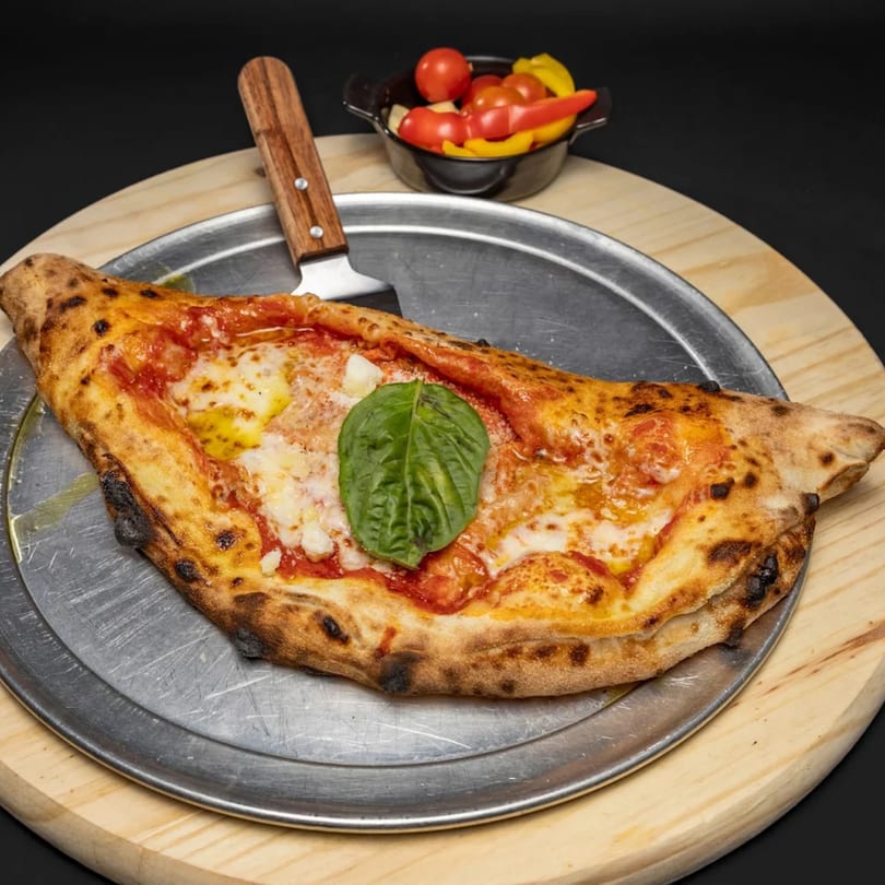 A calzone with melted cheese, tomato sauce, and a basil leaf on a metal pan.